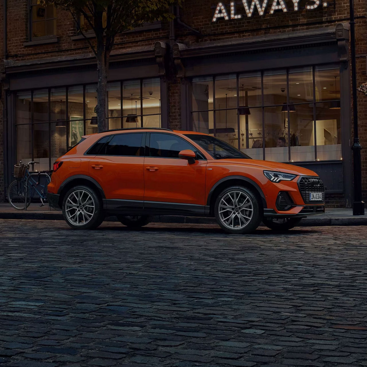 Orange SUV parked on a cobblestone street near brick buildings at dusk.