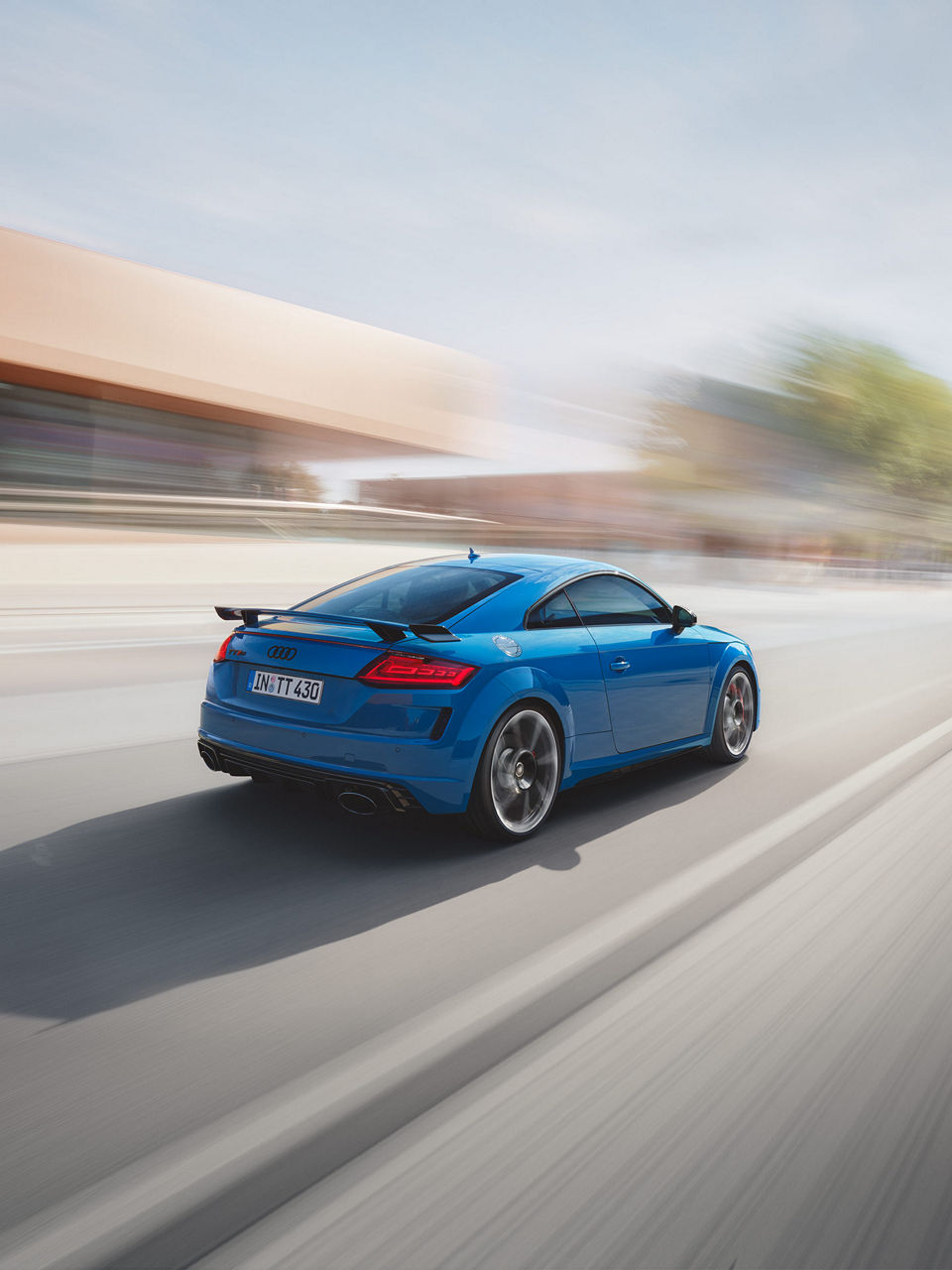 A blue Audi TT RS Coupé speeds along a blurred road, showcasing its sleek design and sporty features with a prominent rear wing.