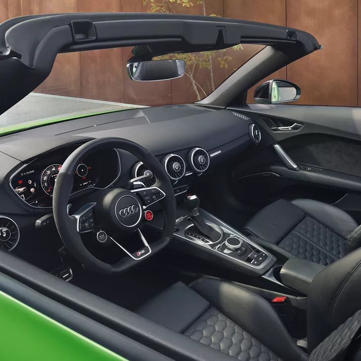 Front seats and cockpit of a green open top Audi TT RS Roadster parked near a wall.