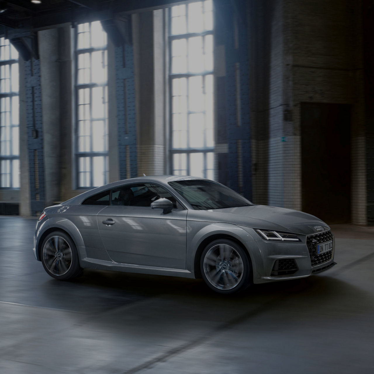 A silver Audi TT parked inside an industrial building with tall windows.