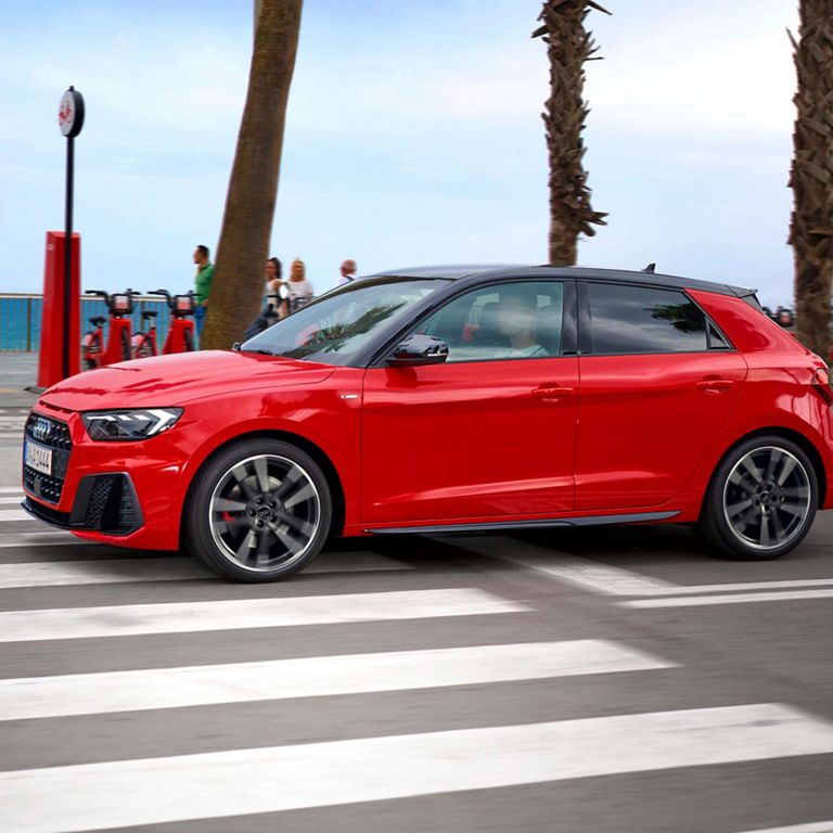 A red Audi A1 Sportback car is driving through a pedestrian crosswalk with palm trees in the background.