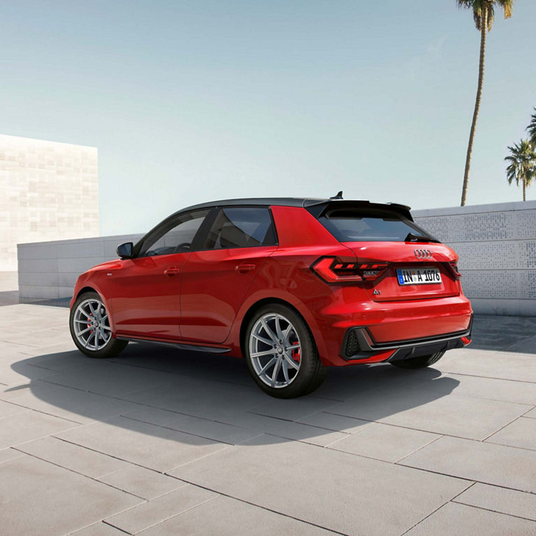 Red Audi A1 Sportback car parked near a wall with palm trees in the background, showcasing a sporty design and alloy wheels.