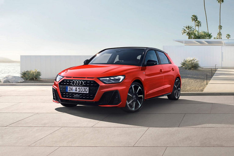 A red Audi A1 Sportback car parked on a paved road near a modern building with palm trees in the background.