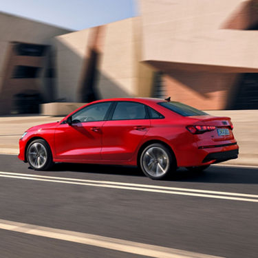 Red sedan driving on a road with modern building in the background.