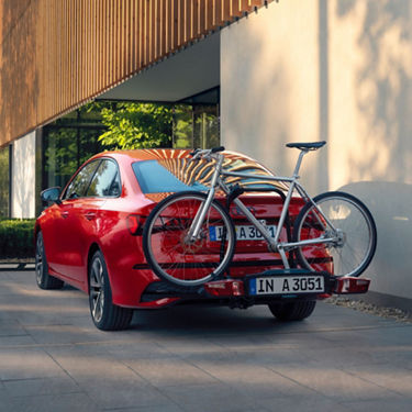 Red car parked with a bicycle rack and bike attached to the rear, in an urban setting.