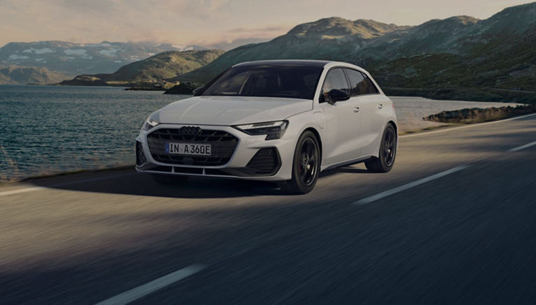 White Audi car driving along a coastal road with mountains in the background.