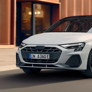 Close-up of a white Audi car's front grille and headlight, with a building in the background.