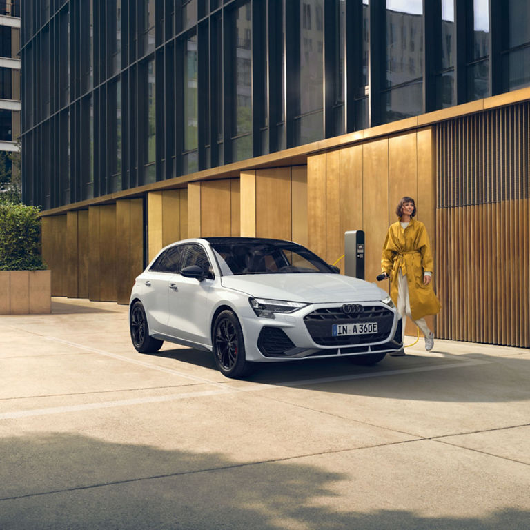 A white Audi electric car being charged by a person in a yellow coat.