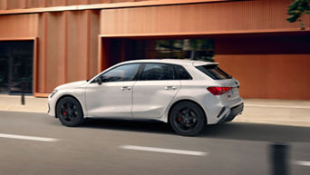 White hatchback car in motion on an urban road with modern buildings.