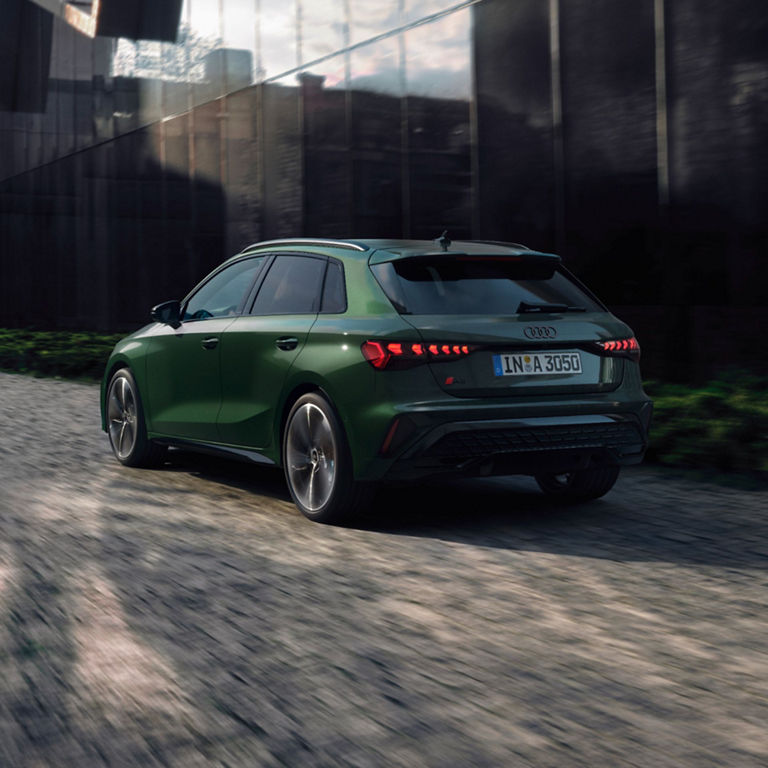 Green Audi car driving on urban street with sunlight flaring in the background.