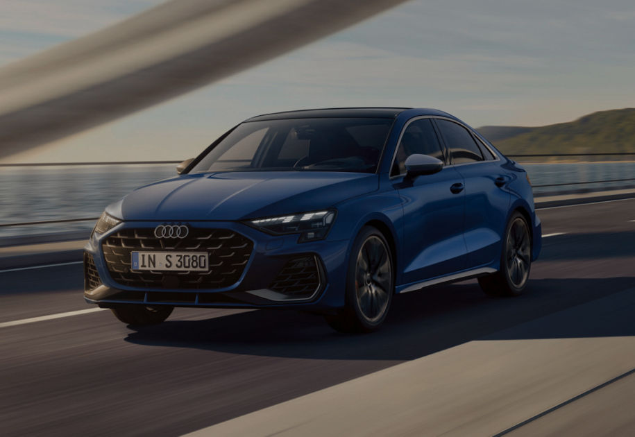 A blue Audi S3 Saloon car driving on a road with a coastal backdrop during dusk.