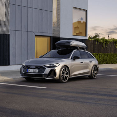 A silver Audi car with a roof box parked outside a modern building at dusk.