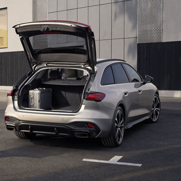 Silver car with open trunk showing a suitcase inside, parked near a modern building.