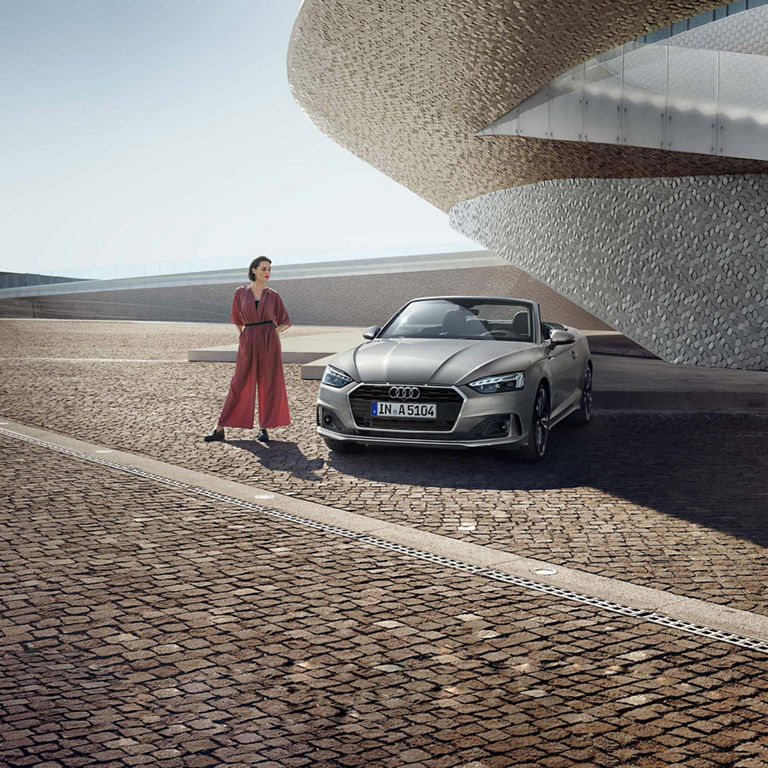 A person in a red dress stands beside a silver Audi car near a futuristic building with a unique texture.