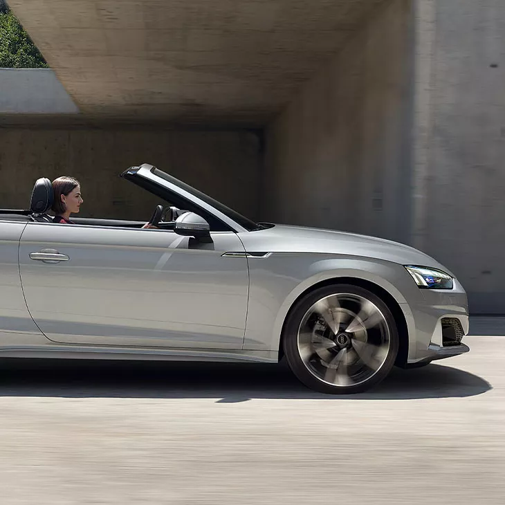 A silver convertible car driving under a bridge. 