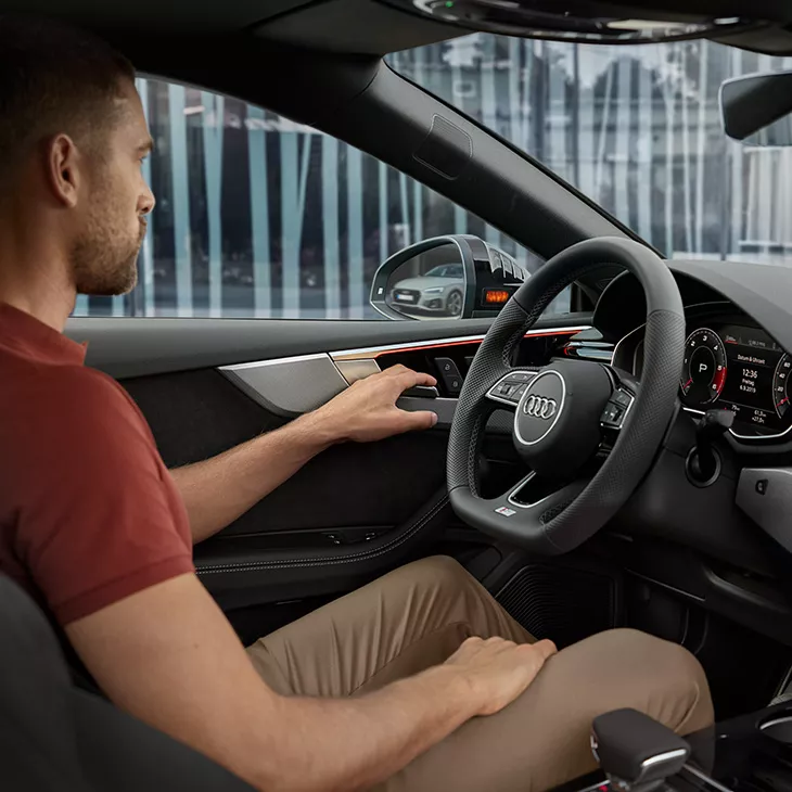 Person sitting in a driver's seat, holding door panel, with a view of the dashboard and Audi steering wheel.