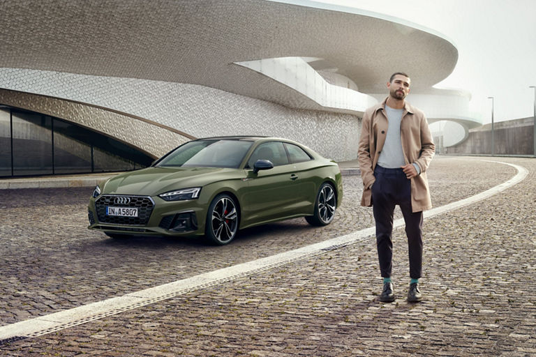 A green Audi car parked beside a modern building with a person standing nearby.