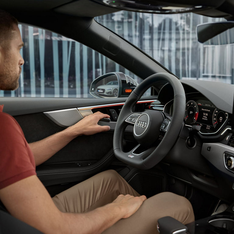 Interior of an Audi car showing the steering wheel, dashboard, and a hand on a control button.