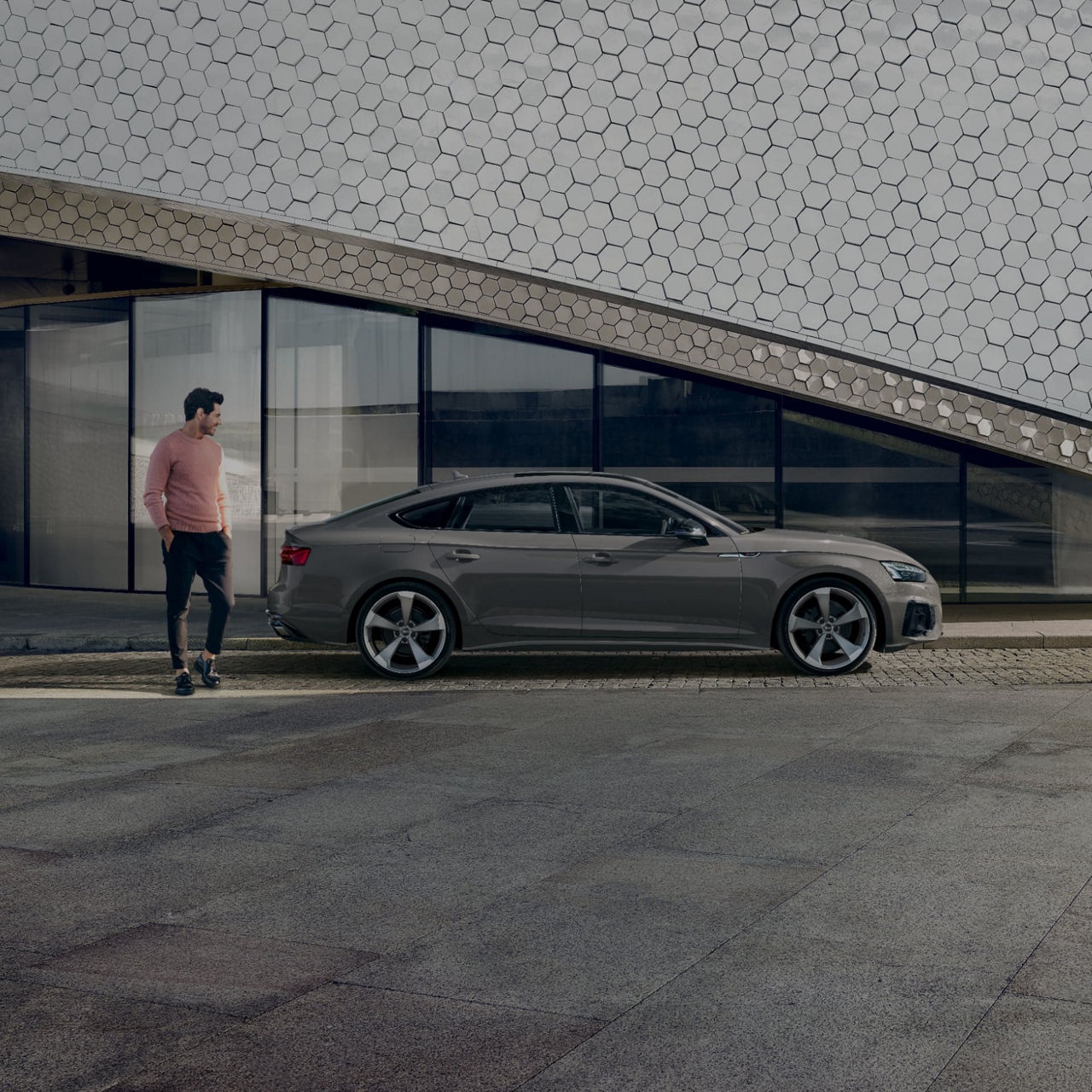 A person walking past a parked silver sedan in front of a building with a hexagonal pattern facade.