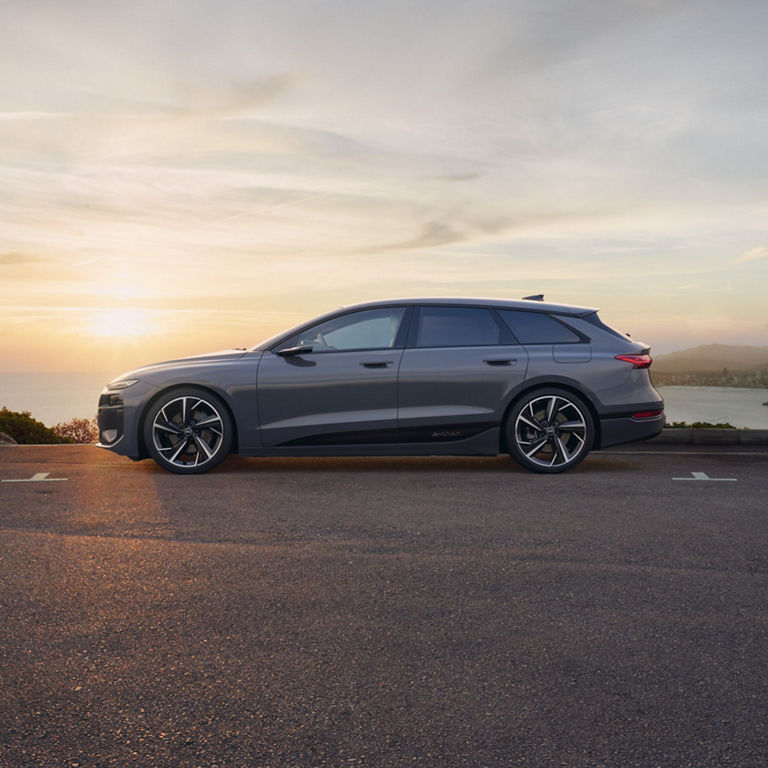 A grey electric car parked by the coast at sunset with city background.