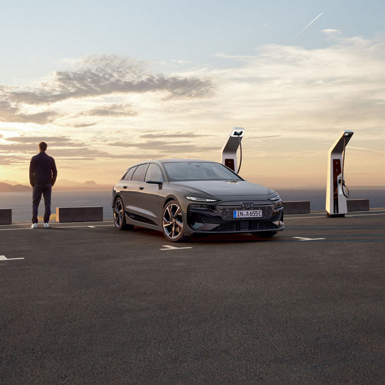 Man standing by an electric Audi at a charging station with a sunset and mountains in the background.