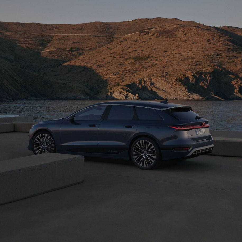 A dark blue car parked near water at dusk with hills in the background.