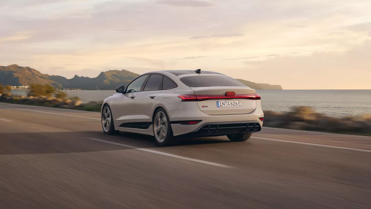 A beige Audi A6 driving on a road alongside a calm sea during sunset.