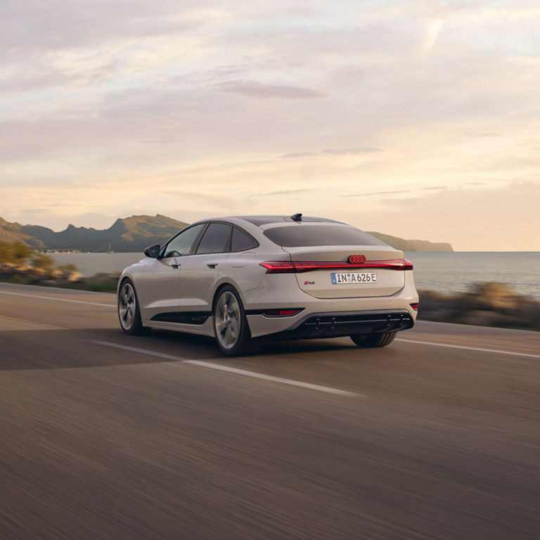 A white Audi sedan driving along a coastal road at sunset.