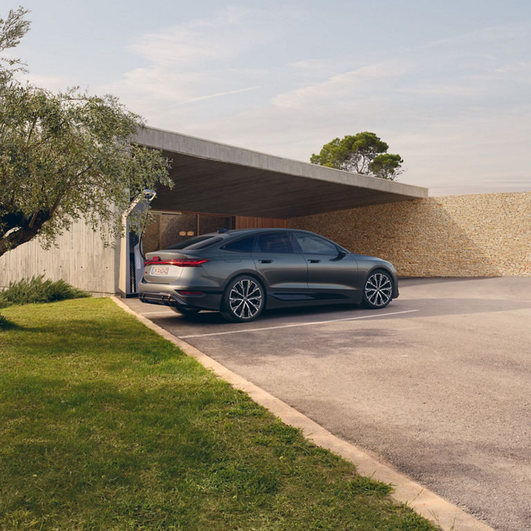 A sleek grey sedan parked outside a modern house under an open garage, near trees and a stone wall.