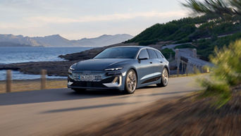 A grey Audi S6 Avant e-tron driving on a coastal road with mountains in the distance.