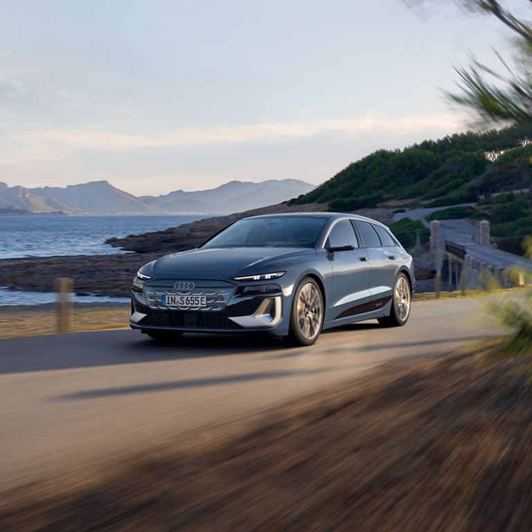 A grey Audi S6 Avant e-tron driving on a coastal road with mountains in the distance.