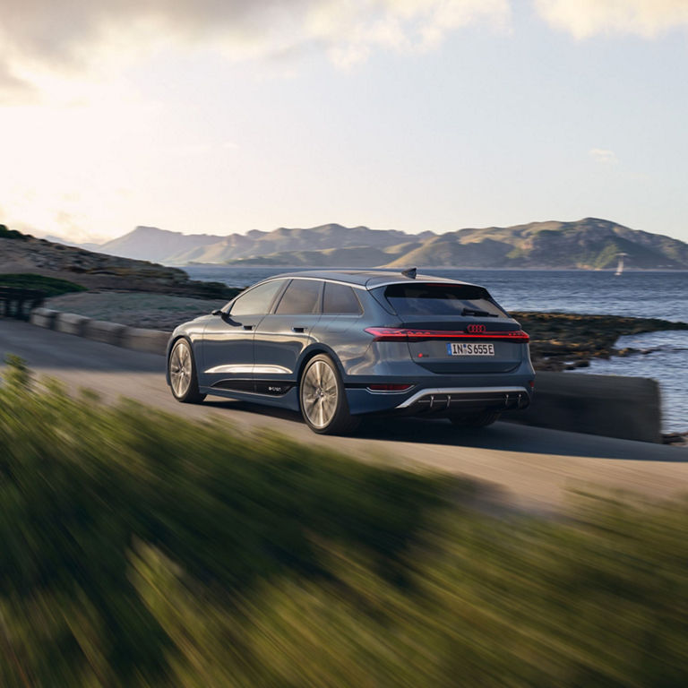 A grey Audi e-tron driving along a coastal road with hills and sea in the background.