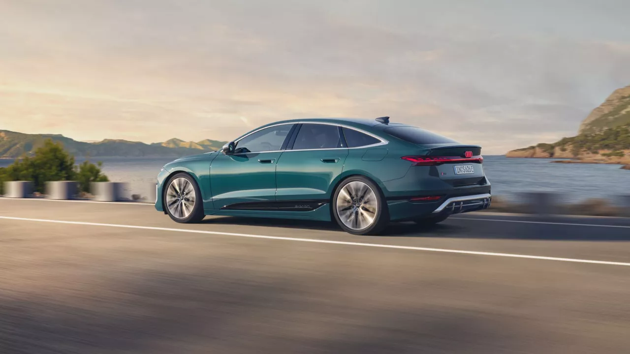 A teal sedan driving along a coastal road with mountains and sea in the background.