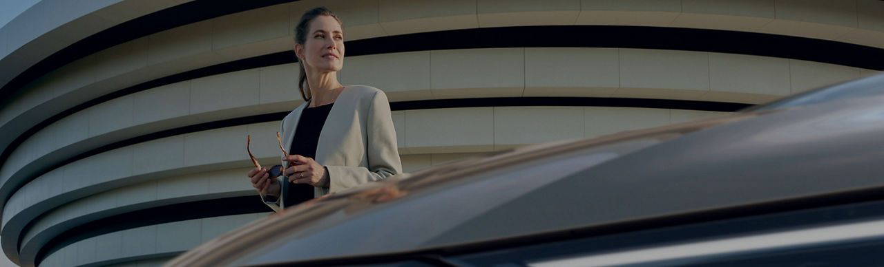 Person in a white top holding sunglasses next to a car with modern building in the background. 