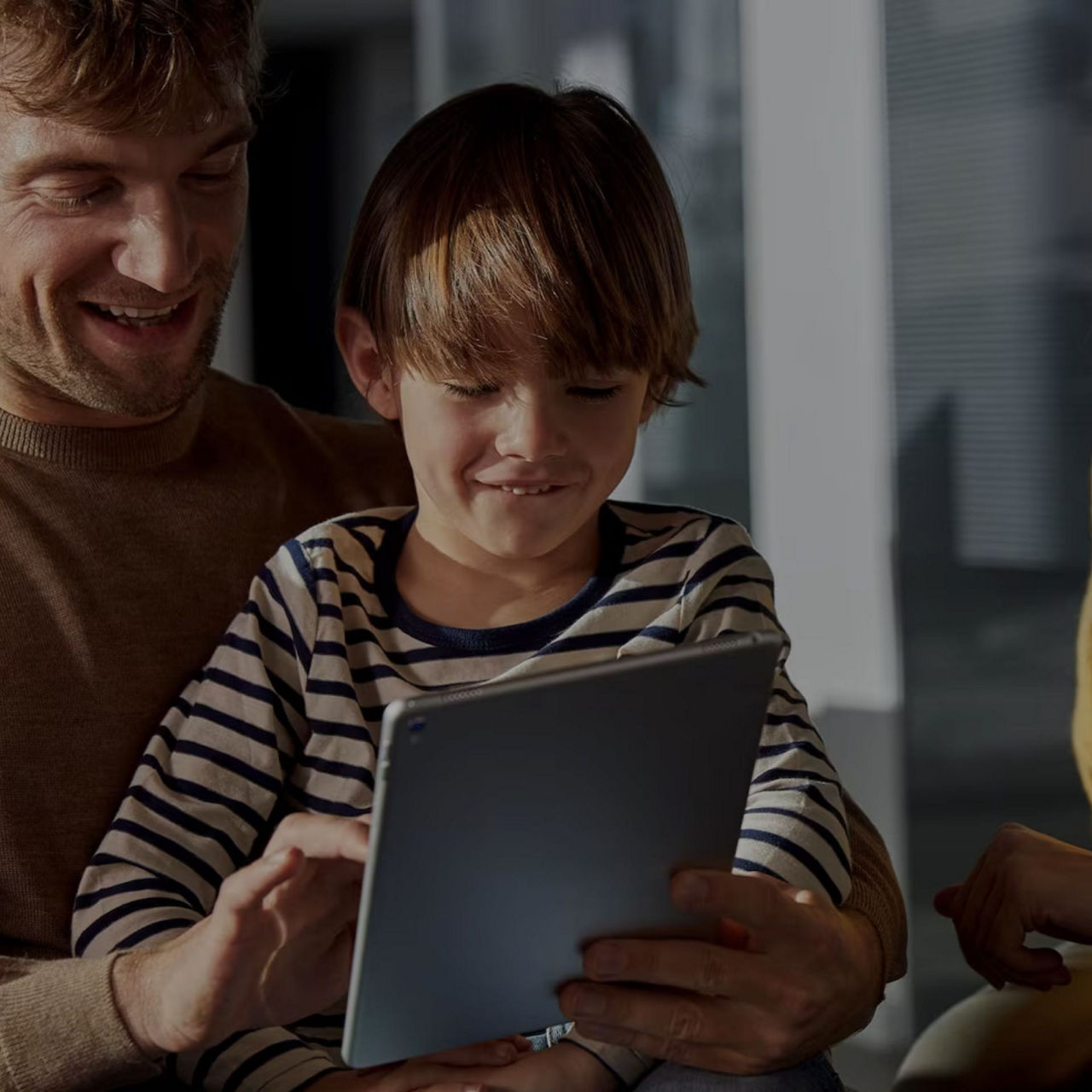 Child in striped shirt holding a tablet with adults on either side. 