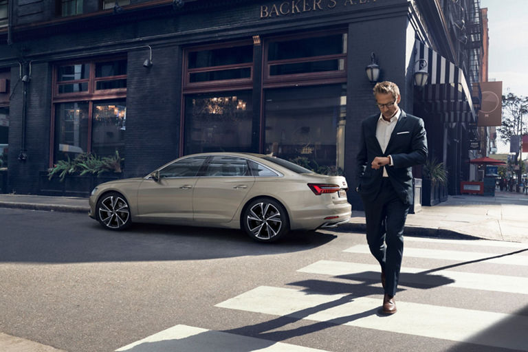A person in a suit crossing a street with a beige sedan car parked on the roadside.