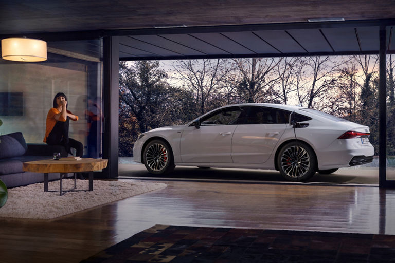 A white sedan parked beside a modern house with large glass windows overlooking trees.