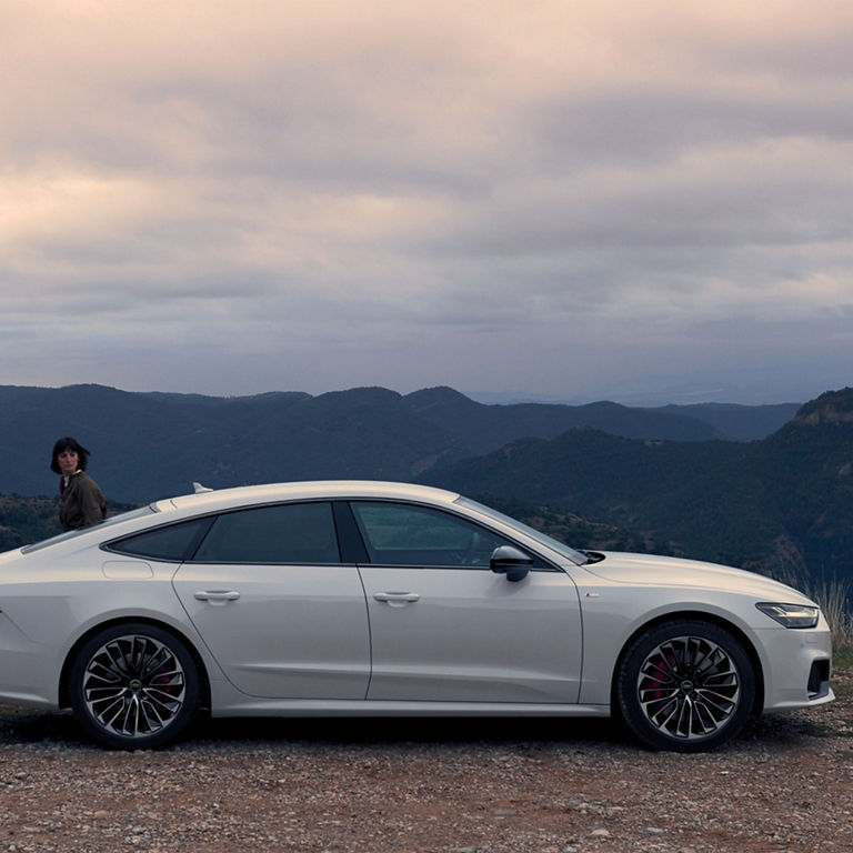 A white sedan parked on a hillside at dusk with mountains in the background.