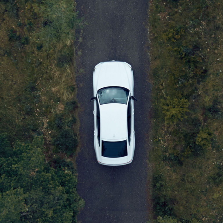 Aerial view of a white car on a narrow road flanked by greenery.