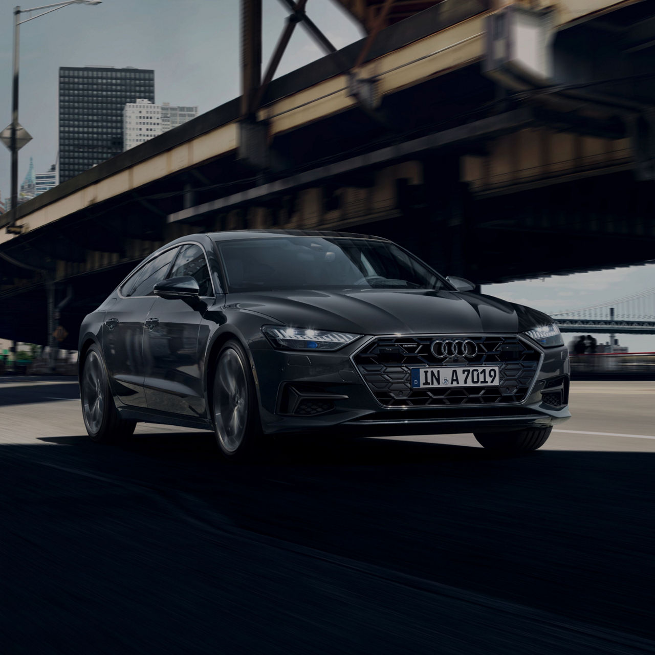 Black Audi driving on city street under bridge, daytime.