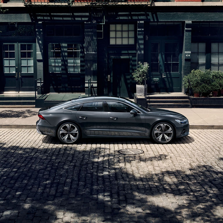 A sedan parked on a cobblestone street by red and grey buildings with balconies.