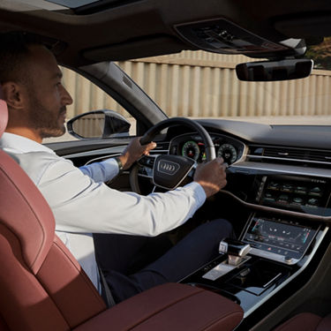 Driver in an Audi vehicle with hands on the steering wheel and dashboard visible.
