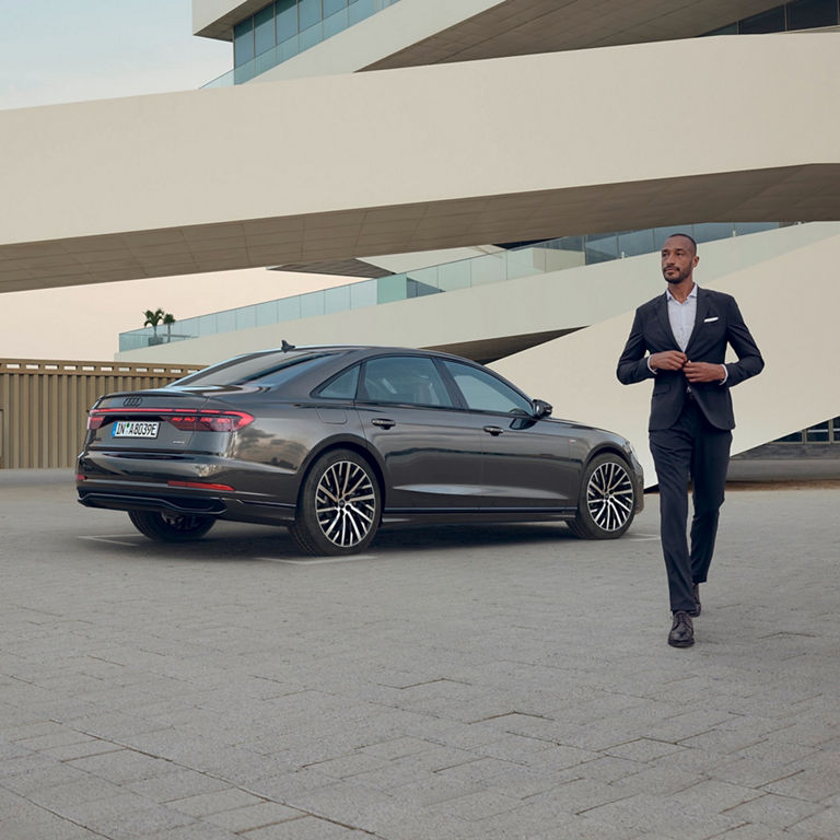 A man in a suit standing beside a dark colour Audi car in a modern architectural setting.