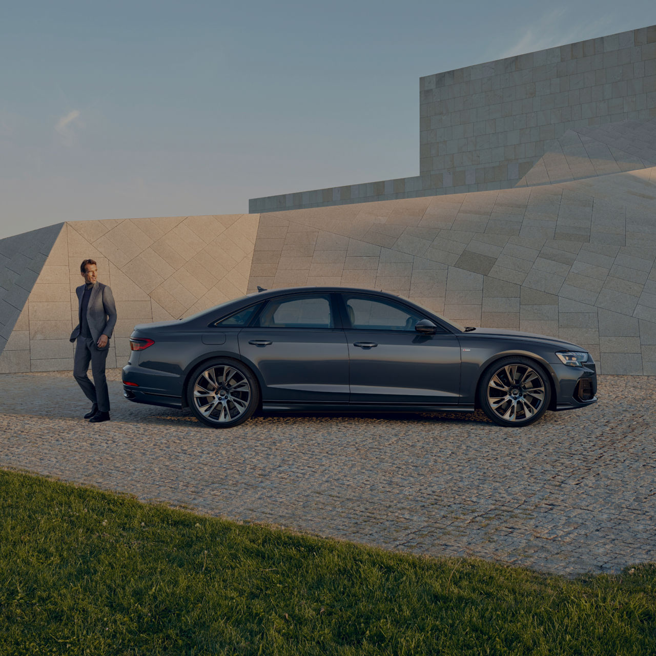 A person in a suit stands beside a sedan near a modern building at dusk.