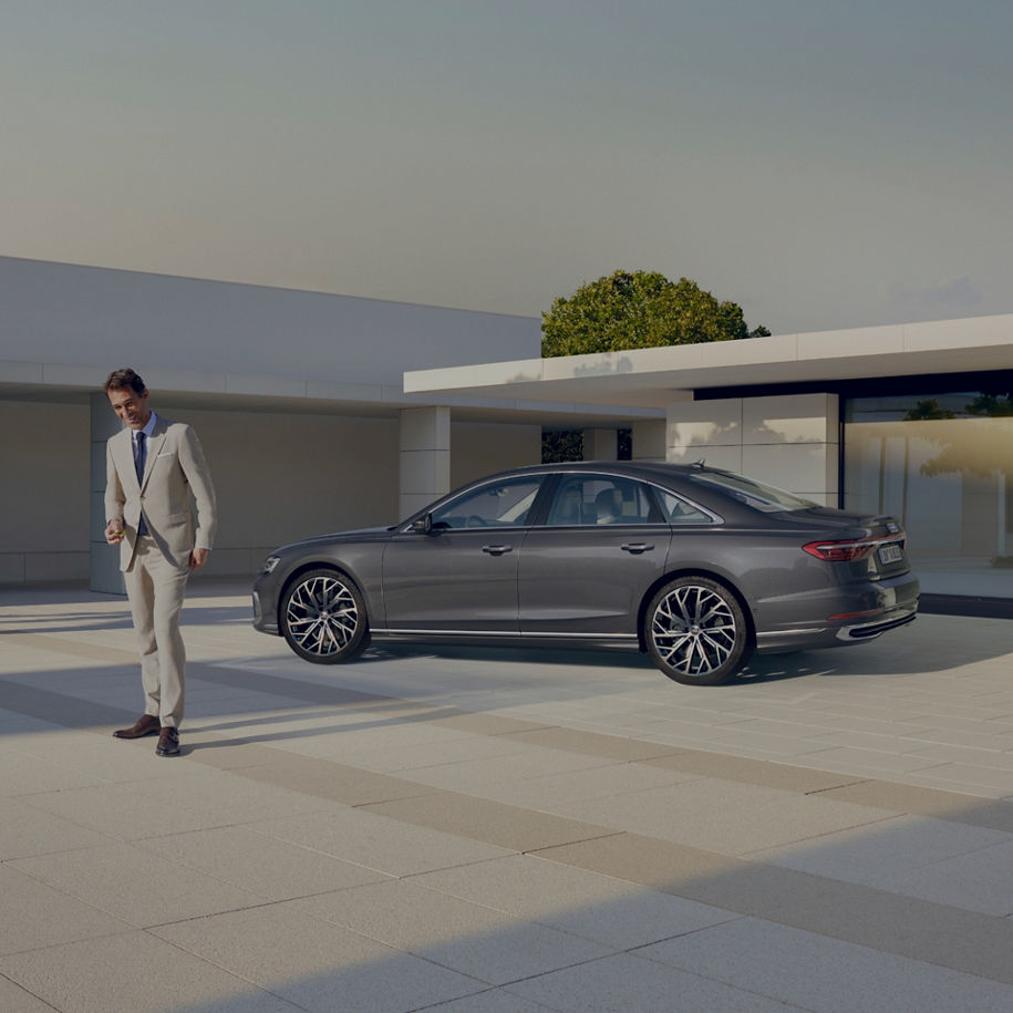 A person in a light suit walking past a parked sedan in a modern driveway setting.