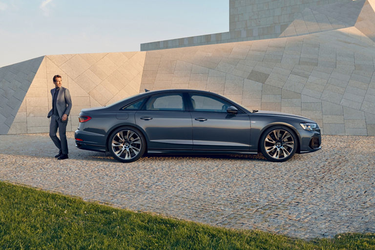 A person in a suit standing next to a sedan on a cobblestone surface with a geometric building backdrop.