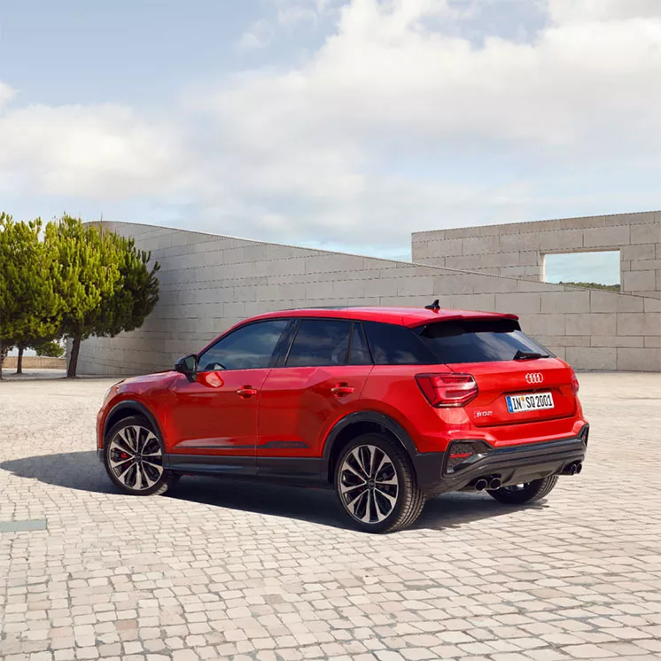 Red Audi SUV parked on cobblestone pavement with trees and modern architecture in the background.