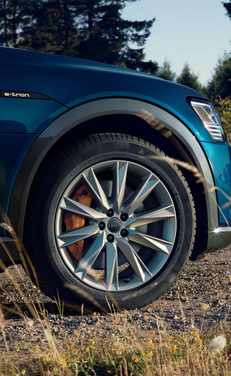 A close-up of a blue car's front wheel and headlight, with "e-tron" badge, on a dirt road surrounded by nature.