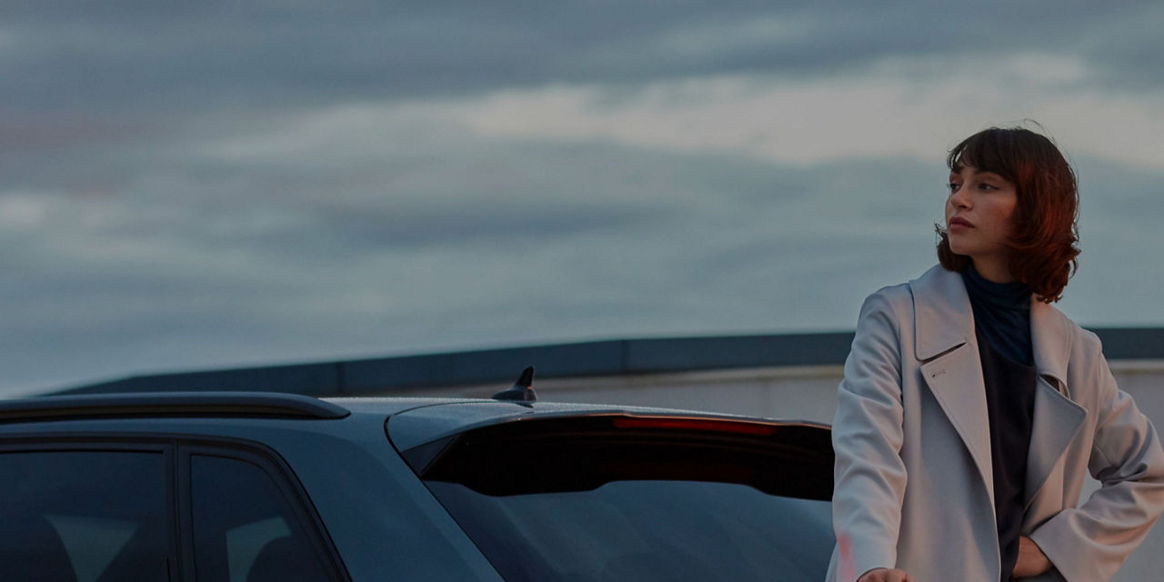 Person in a white jacket leaning on a car roof at dusk.