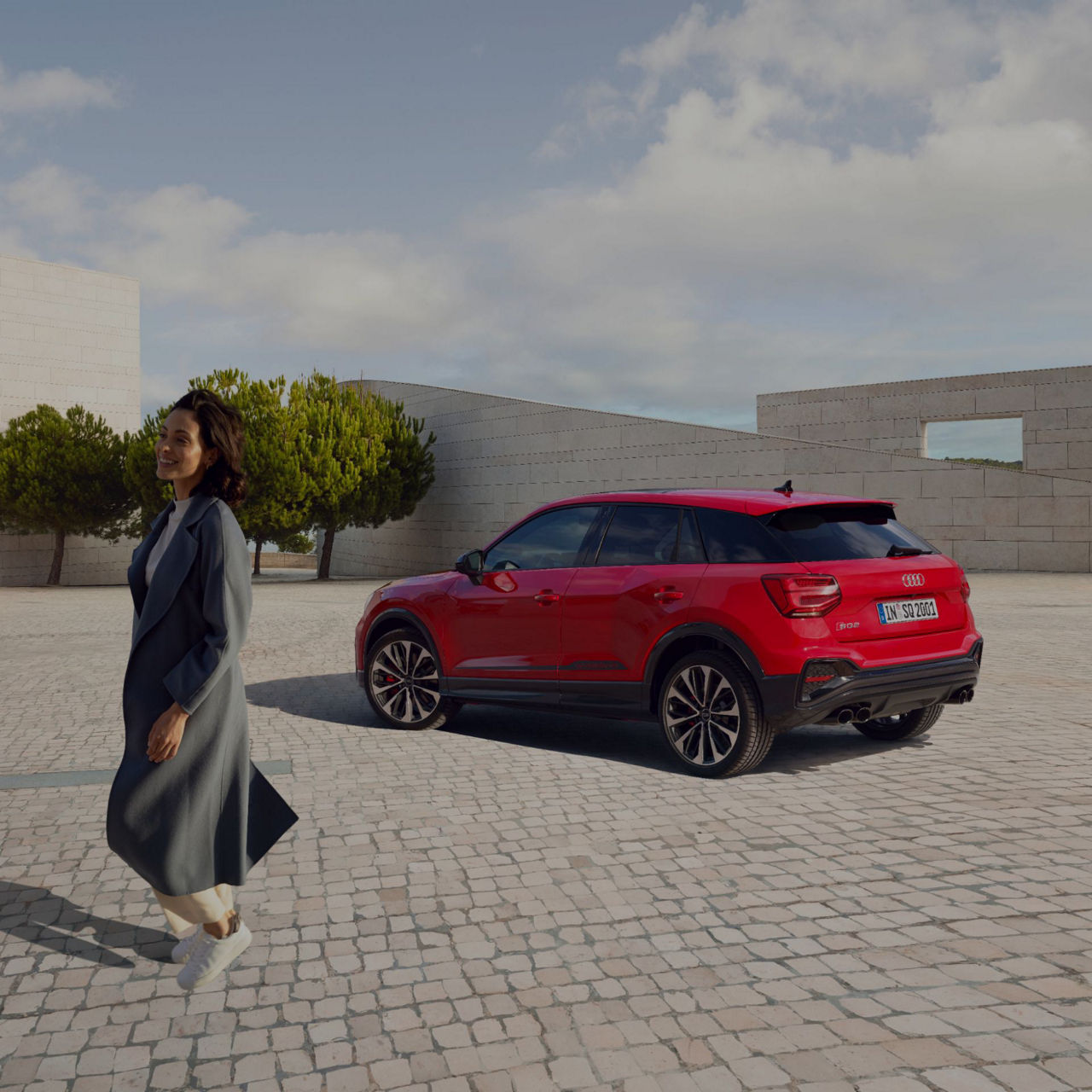 A person walking past a red Audi SUV parked on a cobblestone plaza with trees and a wall in the background.